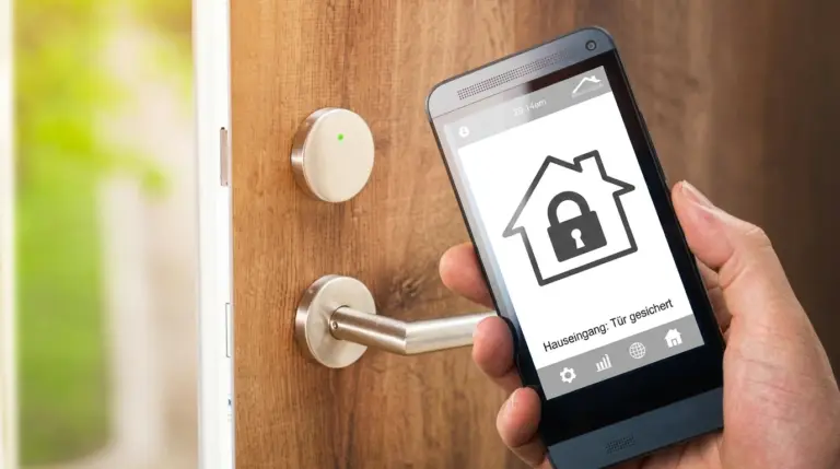 Hand hält ein Smartphone vor einer Holzeingangstür mit moderner Edelstahlklinke und rundem, vernetztem Türsensor, während auf dem Display einer Smart-Home App ein Haus-Symbol mit Vorhängeschloss und der Status „Hauseingang: Tür gesichert“ angezeigt wird.